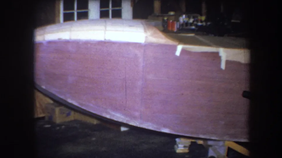 1964 8mm frame from Yucaipa showing the broad front of a boat hull inside a workshop, with tools and materials along the rear surface.