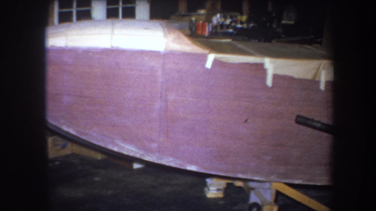 1964 8mm frame showing a reddish-brown wooden boat hull under construction in a dim workshop, with tape and supports visible.