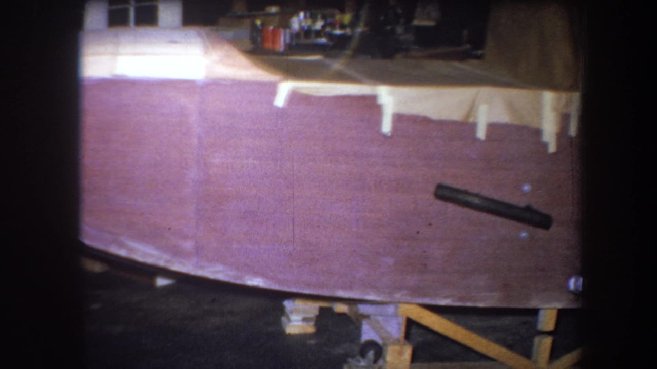 1964 home-movie frame of the wooden boat hull, with taped sections, workshop clutter, and a black cylindrical object along the side.