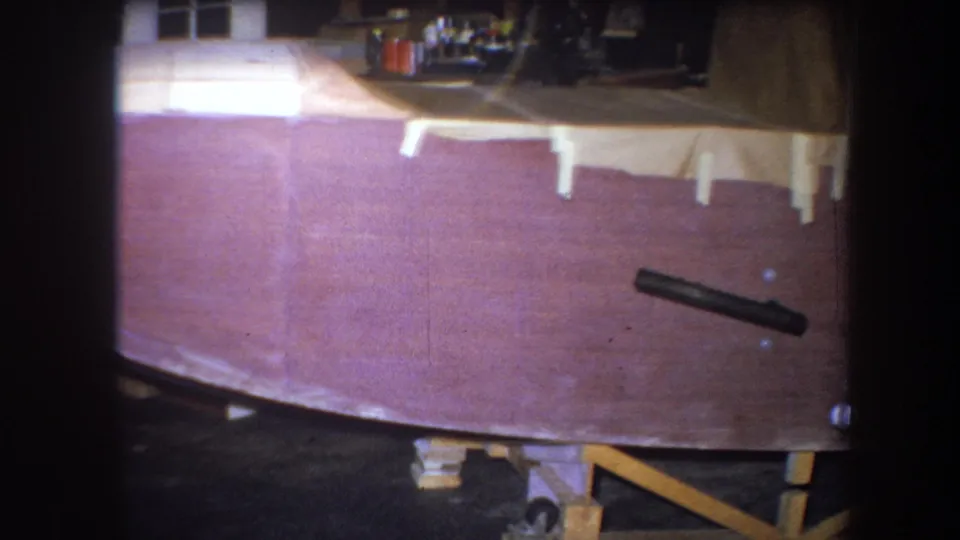 1964 home-movie frame of the wooden boat hull, with taped sections, workshop clutter, and a black cylindrical object along the side.