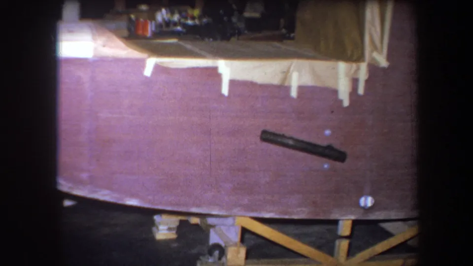 1964 8mm frame showing the unfinished wooden boat hull from a slightly lower angle, with tape tabs and support framing beneath.