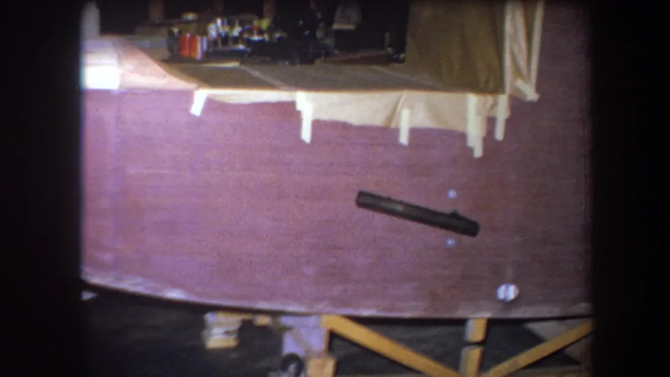 Final 1964 home-movie frame of the wooden boat under construction, showing the hull side, taped areas, and braced support structure.