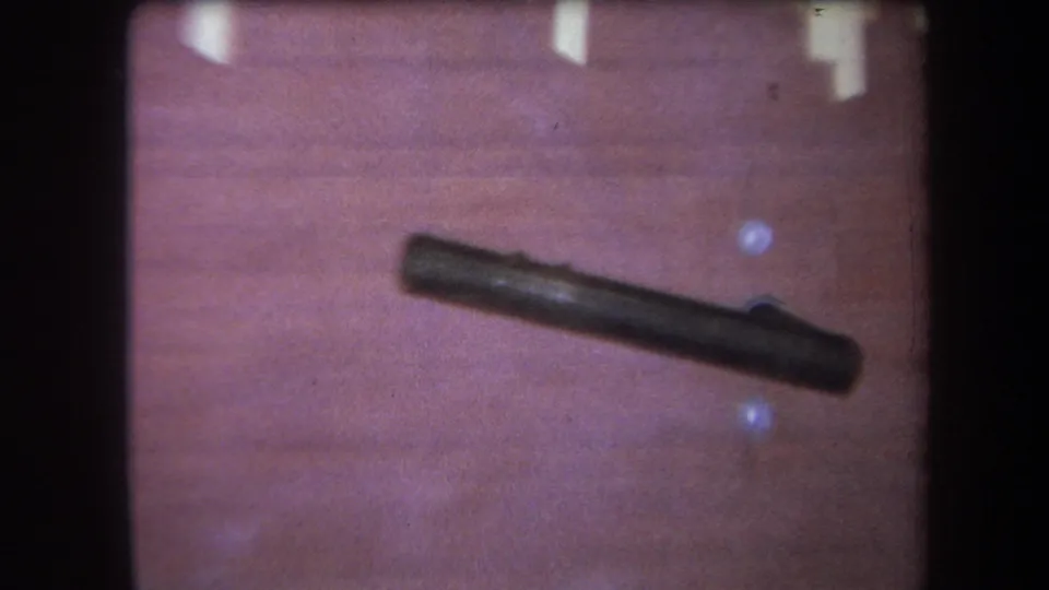 Closer 1964 home-movie view of a dark cylindrical object resting diagonally on a wood-grain tabletop.