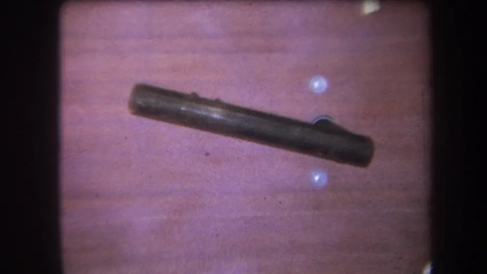 Final close 1964 Yucaipa frame with the dark cylindrical object centered on the wooden table and small bright reflections nearby.