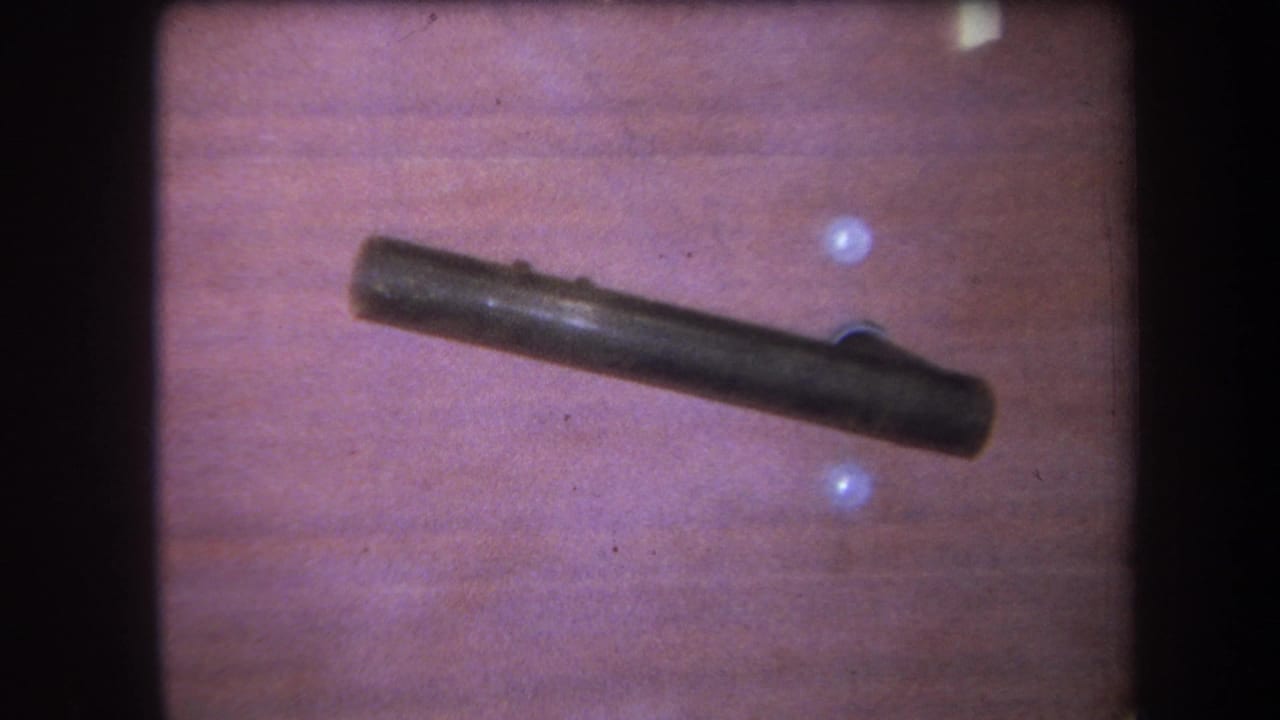1964 Yucaipa home-movie frame showing a dark metal rod or handle against wood paneling with small pale wall holes.