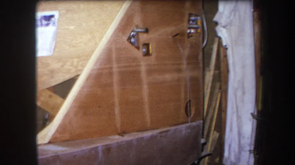 1964 Yucaipa home-movie frame of an unfinished room with wood paneling, metal latches, holes, a hanging cloth, and exposed materials.