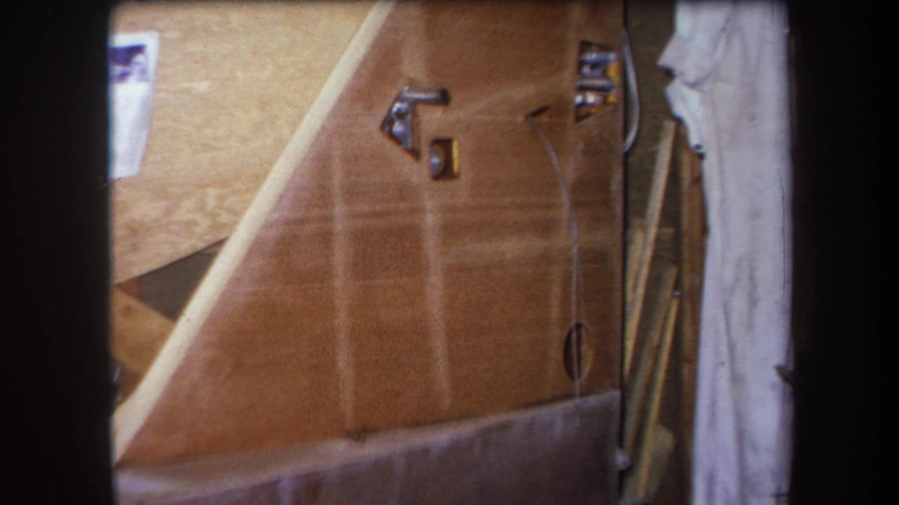 1964 Yucaipa home-movie frame showing a warm wooden surface with metal brackets, a small opening, a thin wire, and cloth along the side.