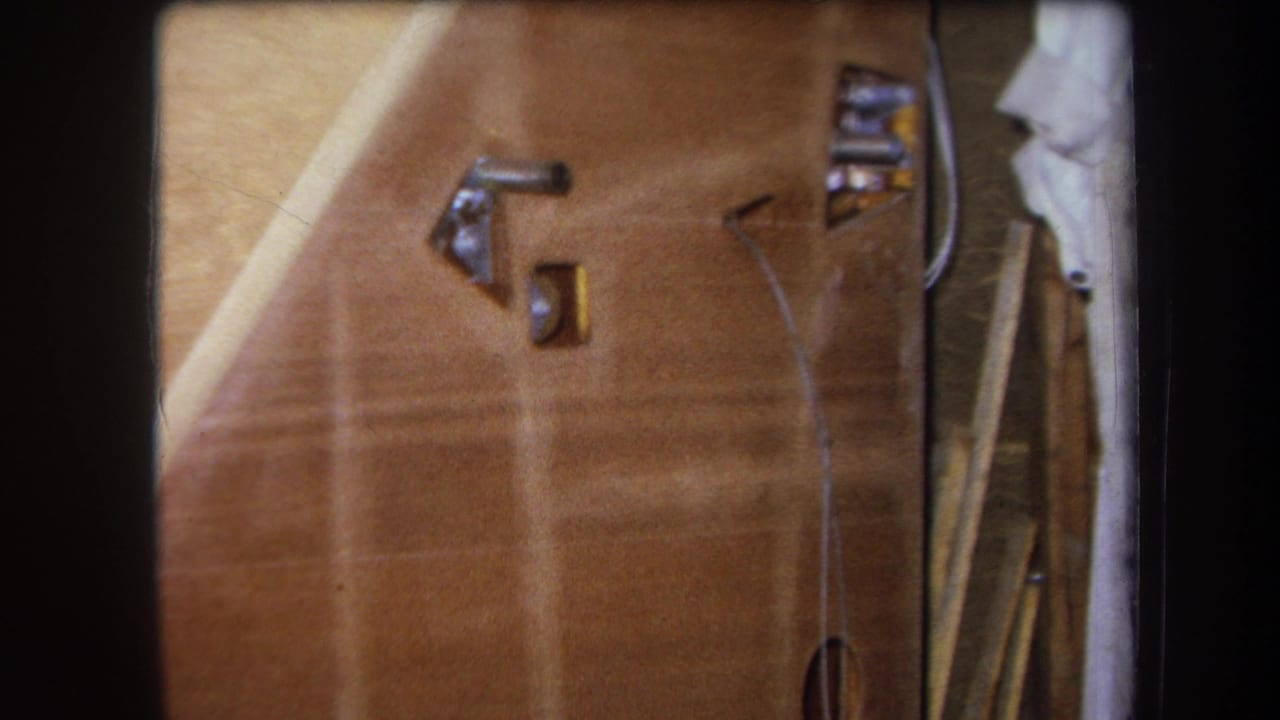 1964 Yucaipa frame with the camera closer to a wooden panel, showing metal hardware, a thin wire, and a pale cloth near the edge.