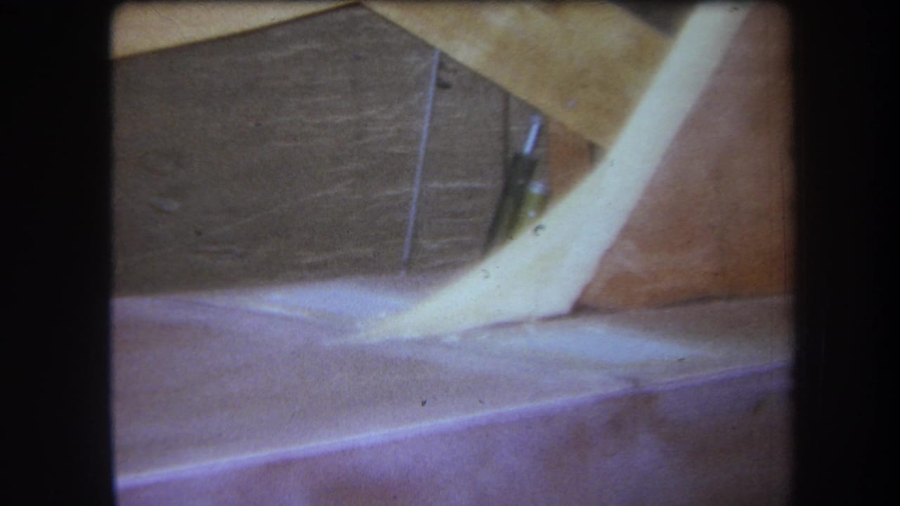 1964 8mm frame from the Yucaipa-area reel showing a dim attic corner with exposed wood, a pale angled form, and soft faded color.