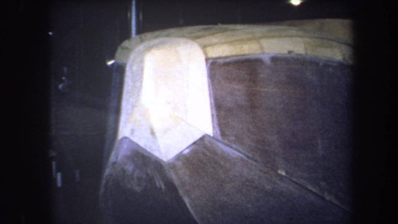 Close 1964 8mm view of a pale wooden aircraft nose section beside dark curved fuselage panels, likely indoors in a workshop or hangar.