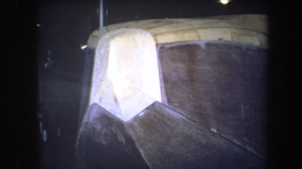 Close 1964 8mm view of a pale wooden aircraft nose section beside dark curved fuselage panels, likely indoors in a workshop or hangar.