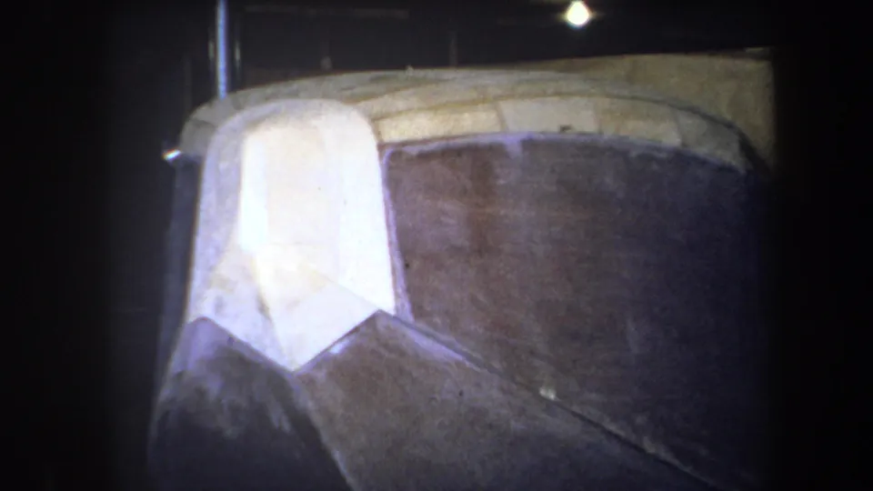 1964 archival frame showing a close-up of aircraft fuselage construction, with a light wooden nose form and dark side panel under workshop lighting.