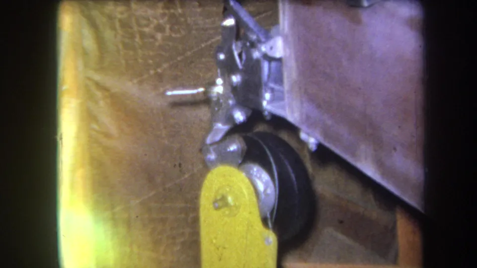 1964 Yucaipa-area 8mm frame showing a small metal bracket and wheel assembly beside a yellow mechanical part.
