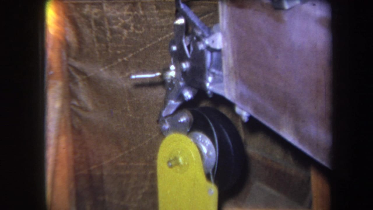 1964 8mm close-up from Yucaipa-area footage showing a yellow control lever, black wheel, and silver linkage against brown textured material.