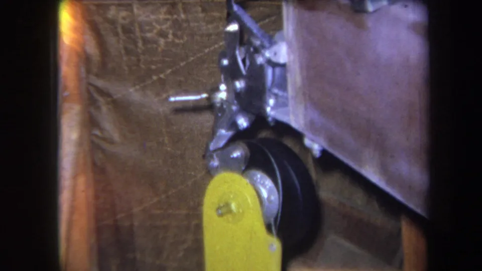 1964 8mm close-up from Yucaipa-area footage showing a yellow control lever, black wheel, and silver linkage against brown textured material.