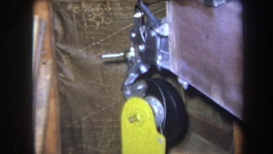 1964 home-movie frame showing the yellow control handle lower in the shot, with black wheel, metal linkage, and brown backing still visible.