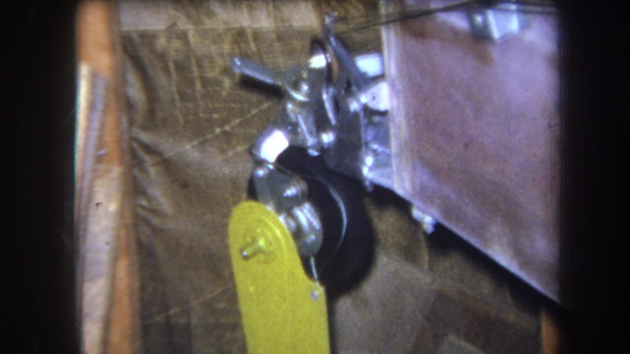 1964 Yucaipa-area 8mm frame showing a pulley-like wheel, metal brackets, wood framing, and a yellow paddle attachment.