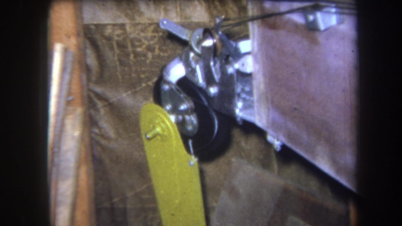 Close 8mm view of a yellow paddle-like part near a black wheel and metal bracketry, supported by the clip's 1964 Yucaipa metadata.