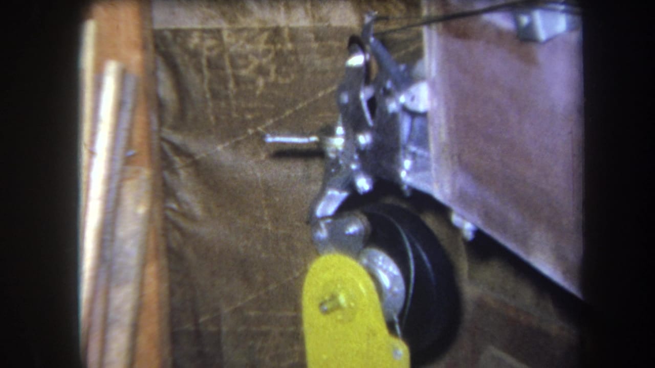 1964 Yucaipa 8mm frame showing a close machine detail with a yellow lever, black wheel, metal bracket, and dark corner framing.