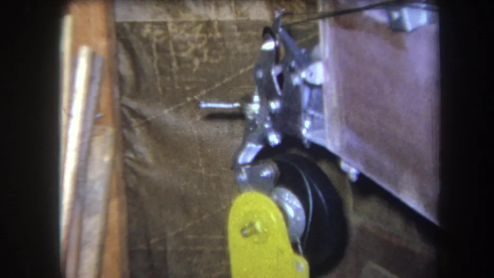 1964 Yucaipa 8mm frame showing a close machine detail with a yellow lever, black wheel, metal bracket, and dark corner framing.