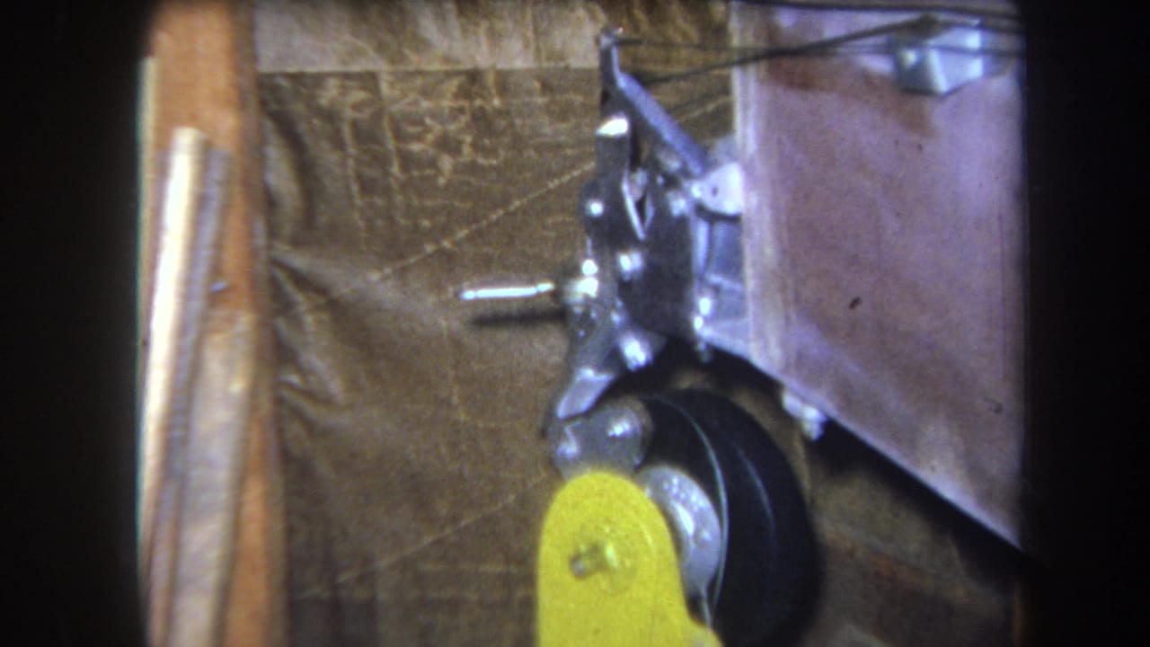 1964 Yucaipa 8mm frame with a static mechanical assembly, yellow lever, black wheel, metal fittings, and brown textured background.