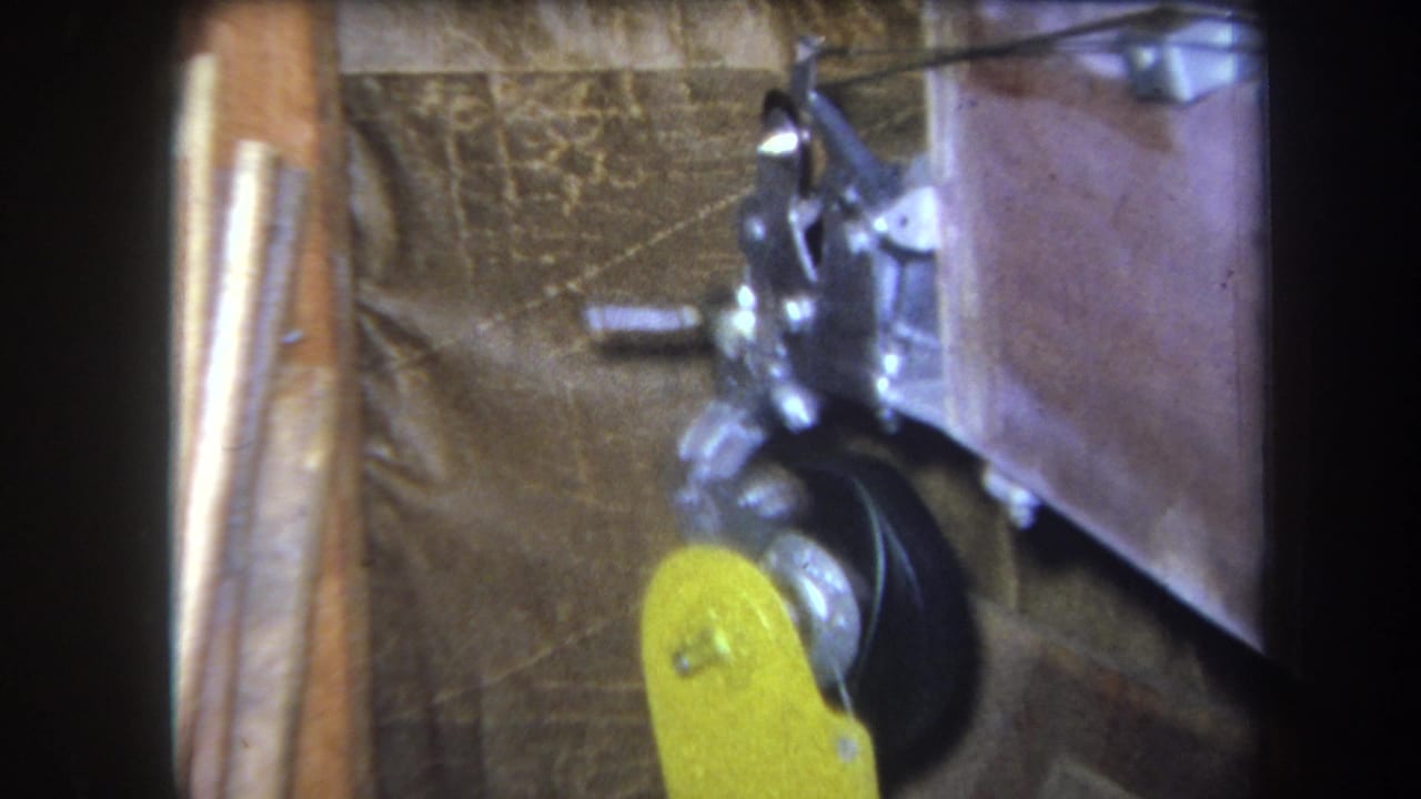 1964 Volmer, Yucaipa 8mm frame showing a yellow lever arm near black rotating parts and silver joints against wood and fabric backing.