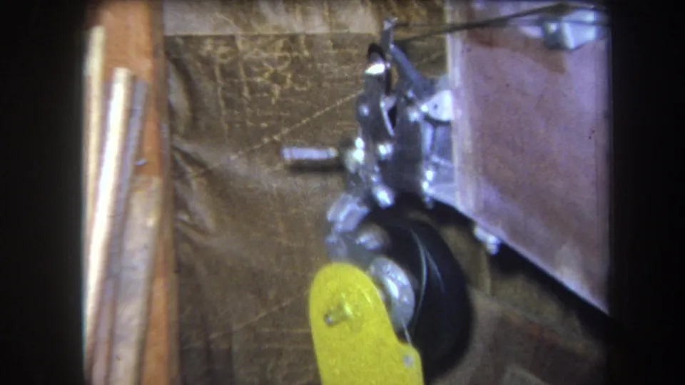 1964 Volmer, Yucaipa 8mm frame showing a yellow lever arm near black rotating parts and silver joints against wood and fabric backing.