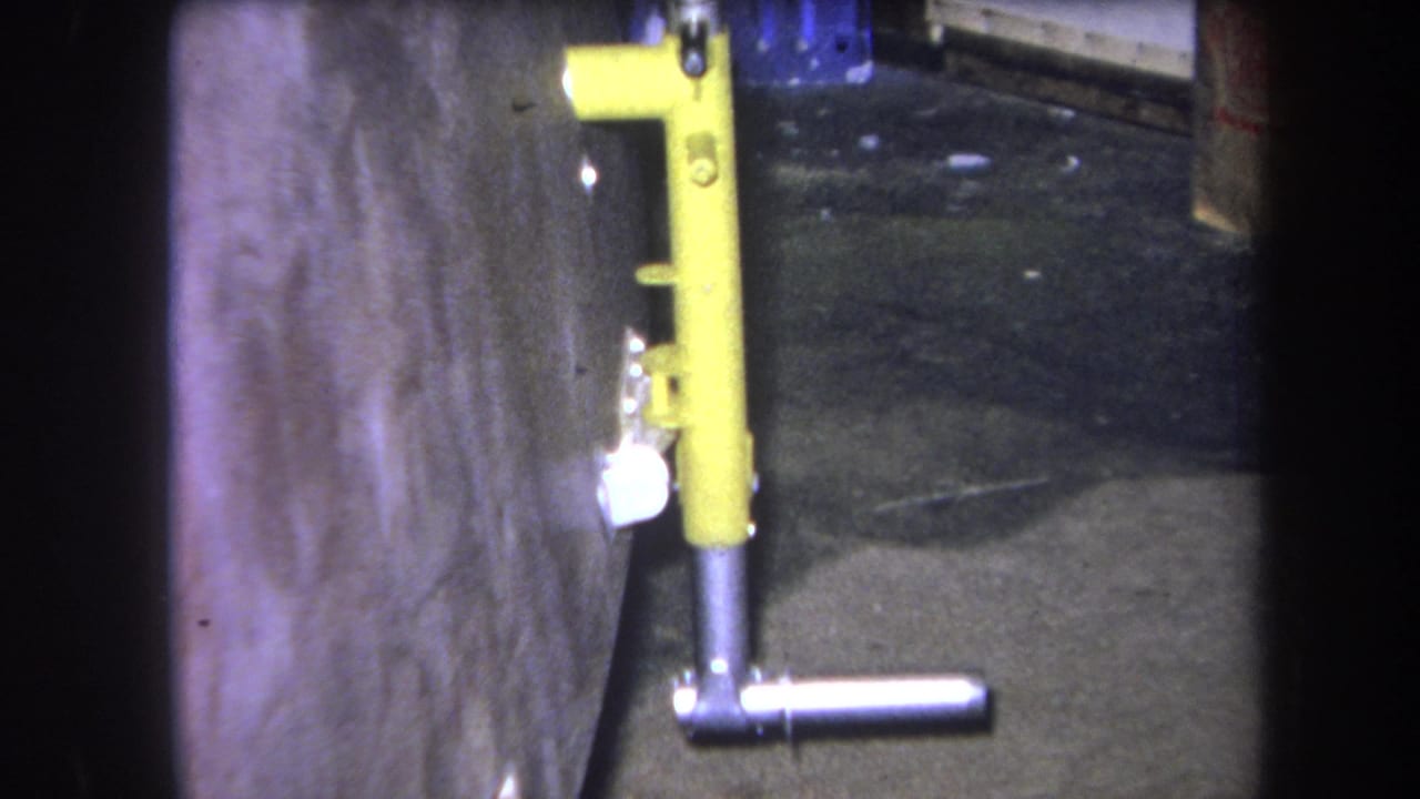 1964 Volmer Yucaipa 8mm frame showing a yellow metal fixture with a silver T-handle beside a concrete wall and dark floor.