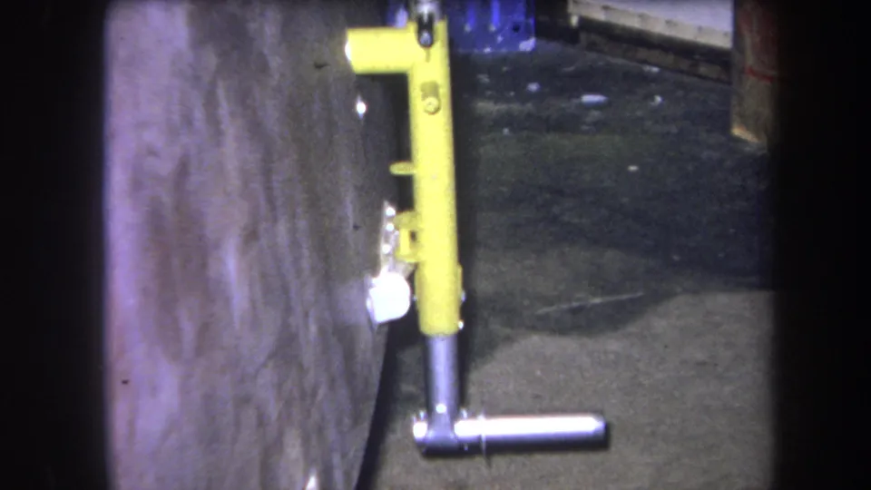 1964 Volmer Yucaipa 8mm frame showing a yellow metal fixture with a silver T-handle beside a concrete wall and dark floor.
