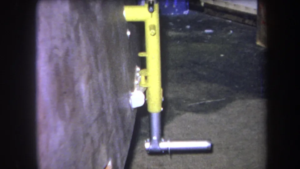 Close 1964 Yucaipa 8mm view of a yellow upright metal part and silver handle near a textured wall and dark floor.