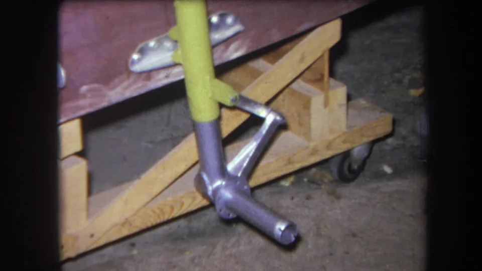 1964 Yucaipa 8mm close-up of a yellow-painted handle, silver mechanical joint, raw lumber support, and wheeled base.