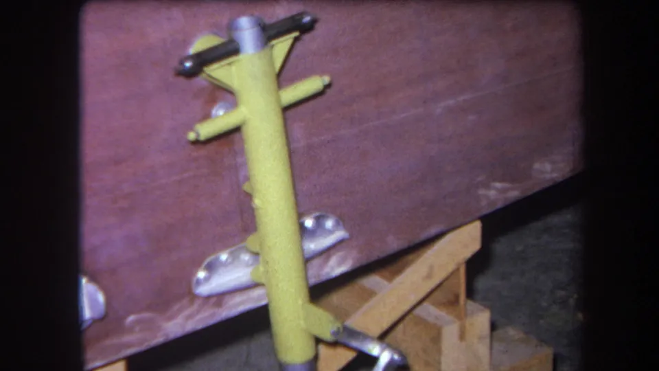 1964 8mm frame of a yellow metal fixture with handle and side arm against a worn wooden workbench or cart.