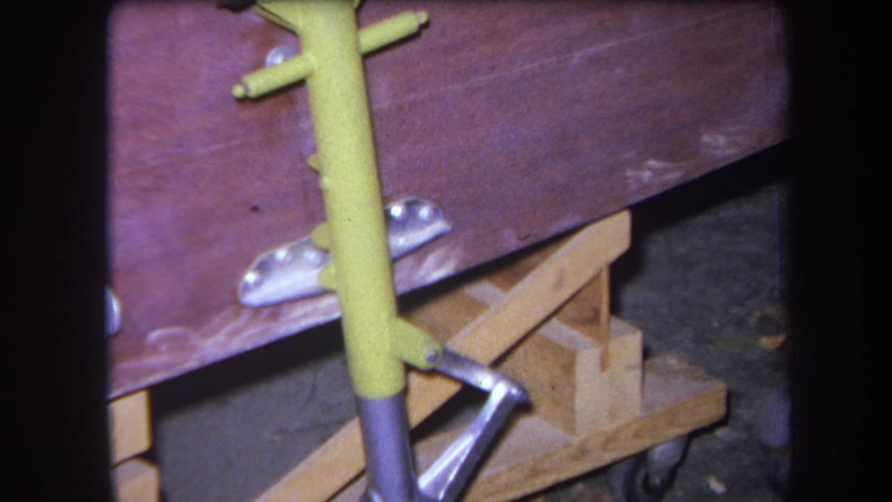 Close view of a wooden cart base with a yellow metal upright, silver bracket, and small caster wheel.
