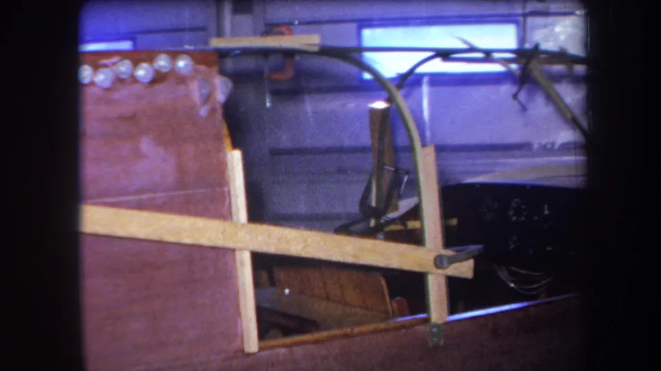1964 8mm frame showing a garage-built vehicle front section with wooden supports, exposed chassis parts, and dim purplish lighting.