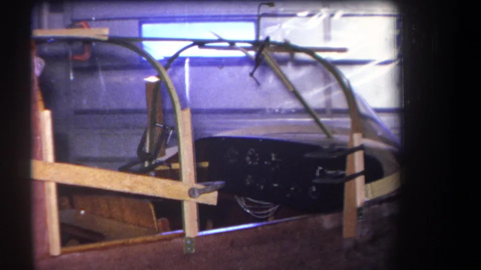 The 1964 home-movie frame records the cockpit assembly with a clear canopy, wooden bracing, and a dark panel of gauges.