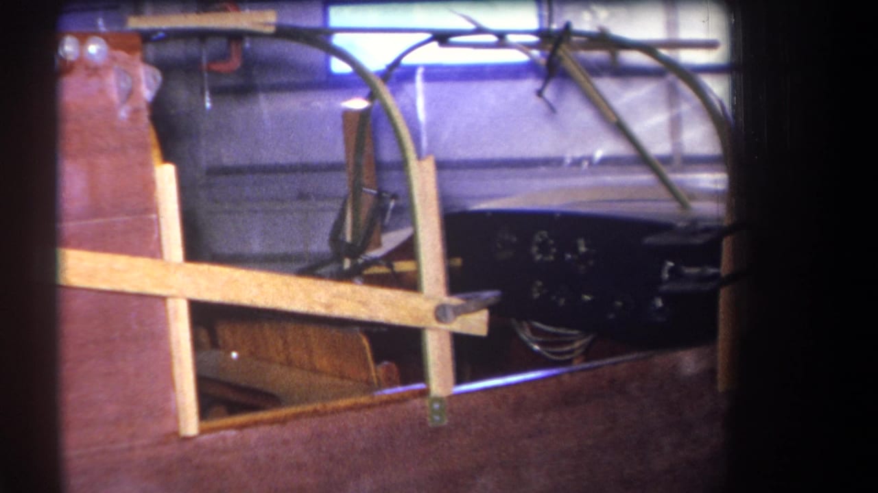 1964 home-movie frame showing the wooden boat cockpit from a slightly shifted angle, with varnished deck, frame supports, gauges, and garage wall.