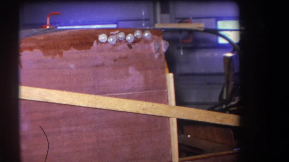 1964 8mm frame from Yucaipa, California, showing a varnished wooden aircraft fuselage section with a diagonal brace and row of silver fasteners.