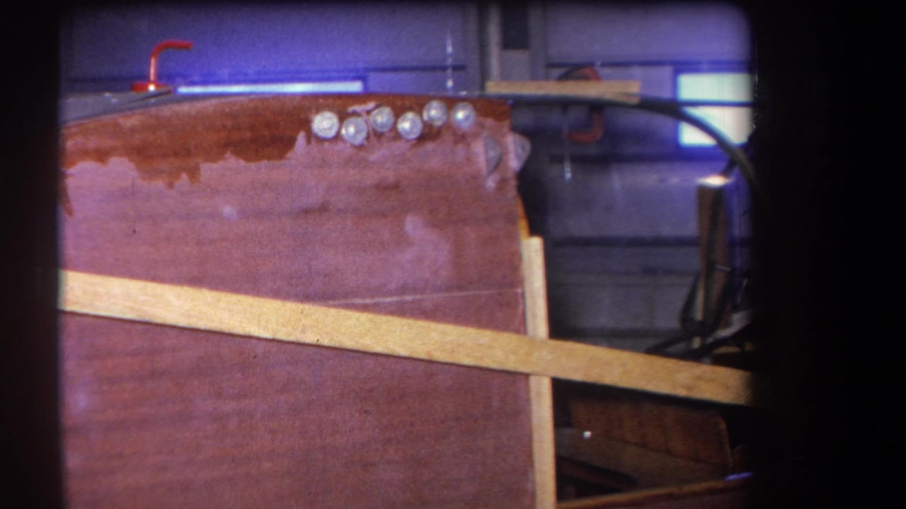 1964 8mm home-movie frame showing an unfinished wooden aircraft fuselage panel, diagonal brace, fasteners, and curved dark cockpit-like framing.