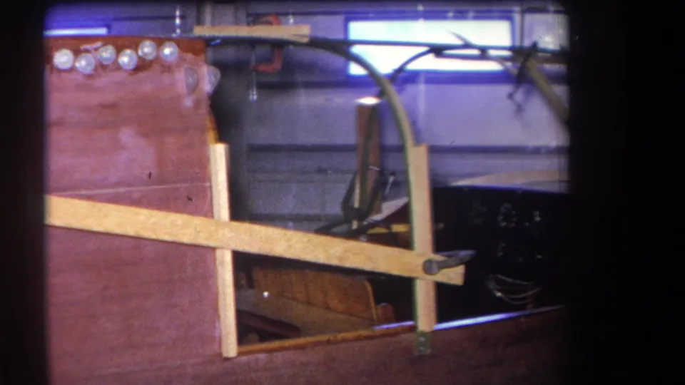 1964 8mm frame from the workshop sequence showing the unfinished fuselage edge, pale bracing, curved metal frame, and dark interior structure.