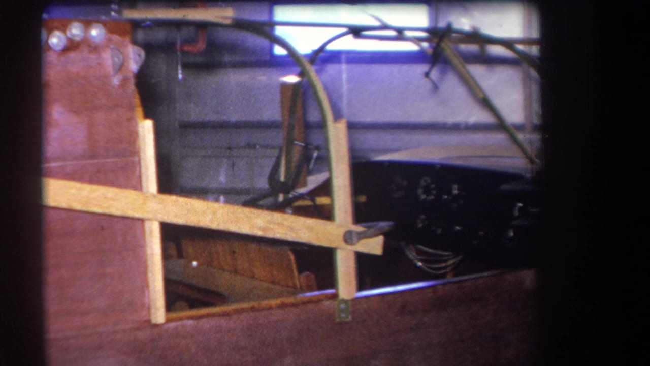 1964 Yucaipa-area home-movie frame showing an unfinished wooden speedboat with braces, canopy, gauges, and wiring inside a garage.
