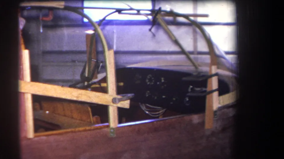 1964 home-movie frame of a wooden speedboat project in a garage, showing the cockpit area, black instrument panel, and temporary supports.