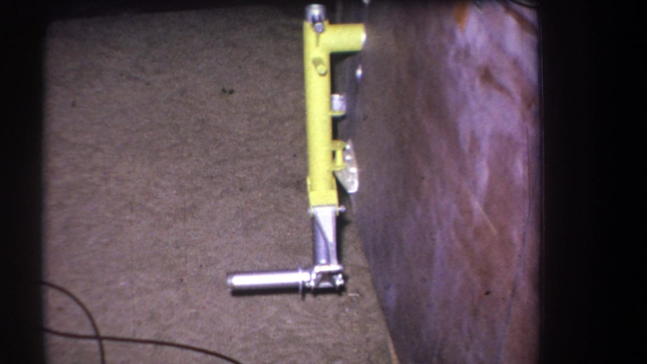 1964 8mm frame from Volmer Yucaipa showing a yellow mechanical lever assembly next to varnished wood and carpeted flooring.