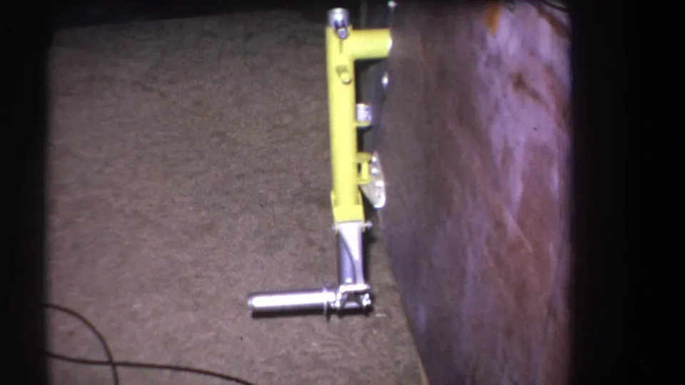 1964 8mm frame from Volmer Yucaipa with a close look at a yellow lever mechanism, metal grip, carpet, and adjacent wood surface.