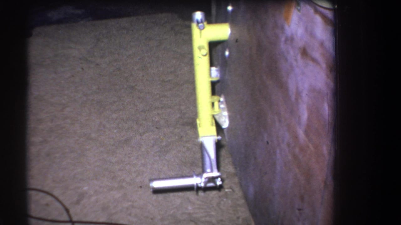 Yellow tool-like mechanical object remains upright beside a wooden structure in grainy 1964 8mm footage from Volmer Yucaipa, California.