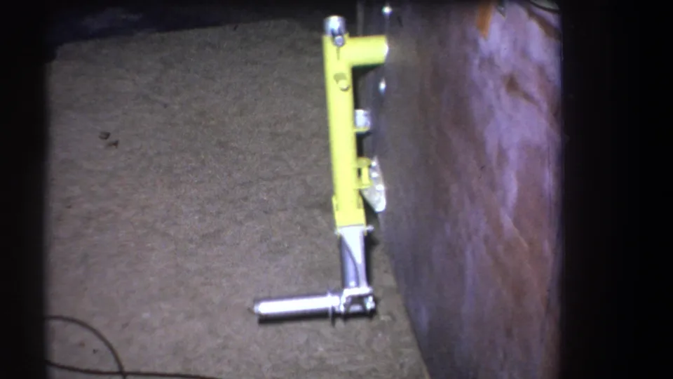 Home-movie frame showing the yellow upright fixture beside a wooden wall, with its metal handle and base visible near the floor.