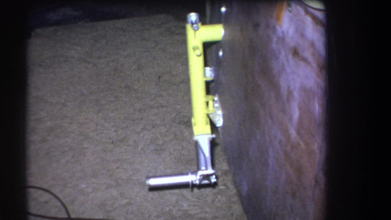 1964 Yucaipa home-movie frame showing a yellow metal lever tool fixed near a wood wall with concrete flooring below.