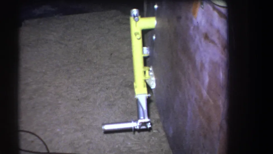 1964 Yucaipa home-movie frame showing a yellow metal lever tool fixed near a wood wall with concrete flooring below.