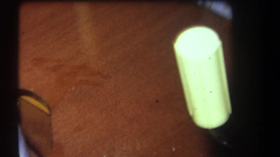 1964 8mm close-up with a pale yellow cylinder standing near varnished wood and partial metal or dark hardware.