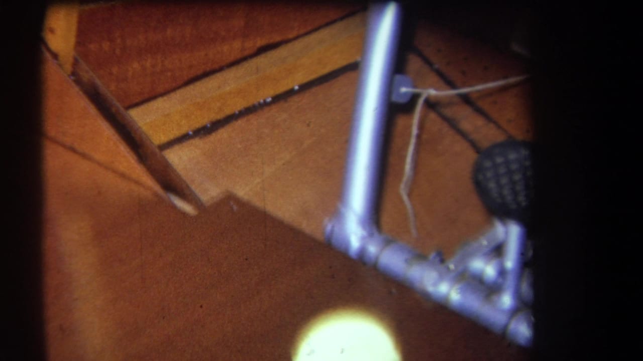 1964 8mm frame showing a small aircraft cockpit floor, silver control stick, wiring, and foot pedal in Yucaipa-area home-movie footage.