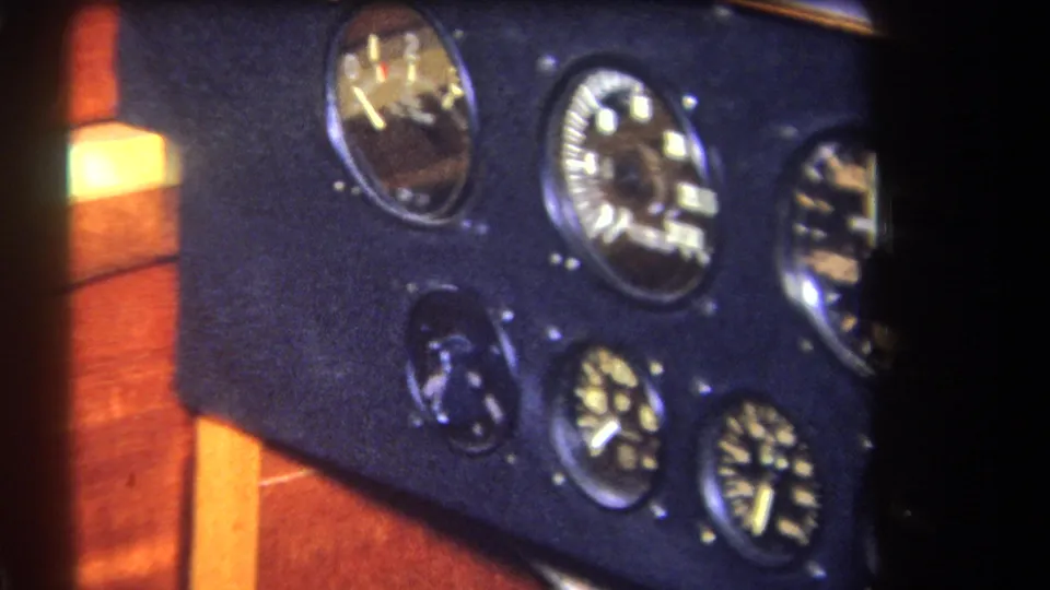 1964 8mm frame showing a tilted view of aircraft gauges, a dark instrument panel, and orange-brown wood cockpit surfaces.